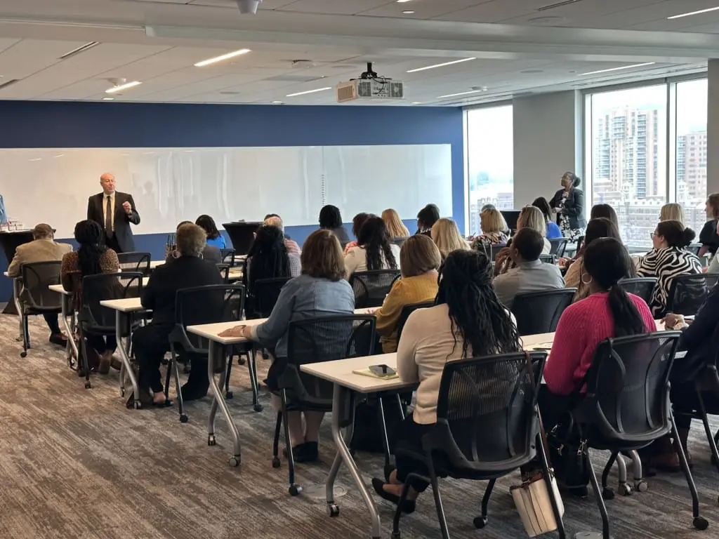 A wide shot shows a man in a suit and tie standing at the front of a classroom, speaking to an audience of approximately 30 people seated at individual desks. The man is balding and has a light complexion. The classroom features a large white board behind him and large windows on the right side of the frame, offering a view of other buildings. Most of the audience members are seen from behind, and they appear to be primarily women of diverse ethnicities. The room has light-colored carpet with a geometric pattern.