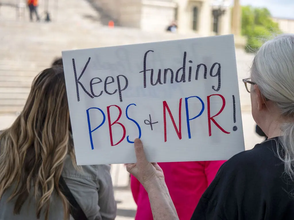 A protester holds a sign in support of funding for public media during a May 1, 2025, rally at the Kansas Statehouse in Topeka as part of a 50501 national day of action. (Sherman Smith/Kansas Reflector)