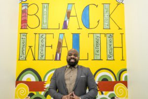 Brandon Calloway, Director Ejecutivo y Cofundador de Kansas City G.I.F.T.. posando delante de una obra de arte que incluye las palabras "BLACK WEALTH".