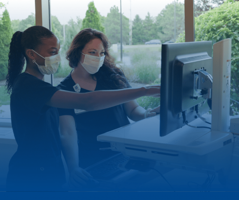 Two women with Covid masks on examine a monitor in a work environment.
