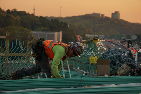 Male construction worker working on a roof during sunset. A city can be seen in the background.